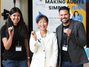 Michael Bryant, Alex Baron, Shaowen Hua smiling, holding LEGO figures. Background shows a sign reading "Making Audits Simple & Fun." Bright, joyful setting.