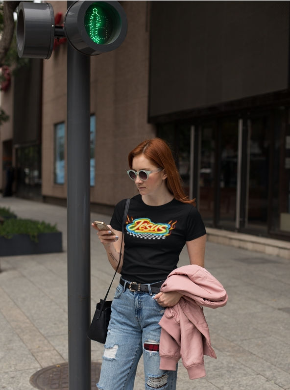Woman wearing a black graphic t-shirt with colorful “Jesus” flame design, standing on a city sidewalk while looking at her ph