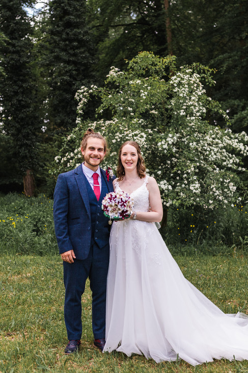 Portrait photograph of bride and groom with arms round each other, smiling at the camera on their wedding day at Stourton Estates in Horncastle. Groom is in a. blue suit and red tie whilst bride is in a ivory floor length gown