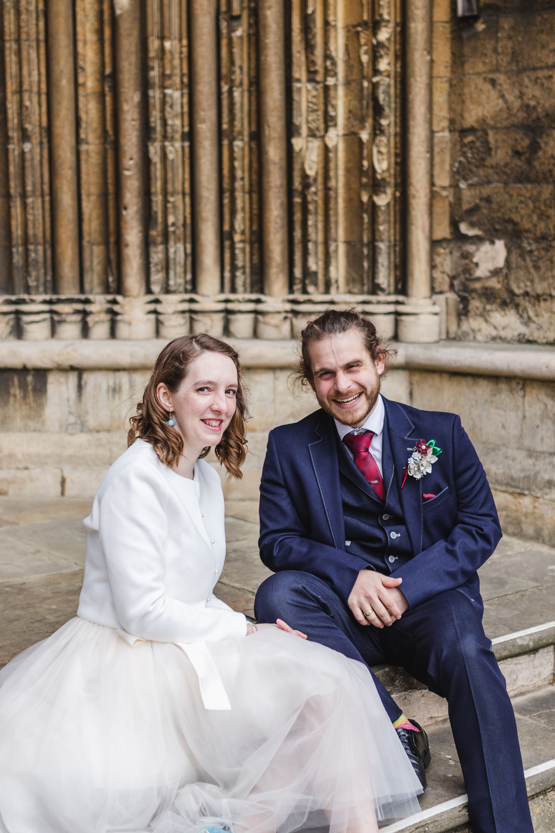 Bride and groom laughing at camera as they sit on steps outside Lincoln Cathedral on wedding day
