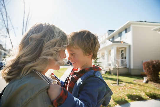 The image shows a mom with her young son standing outside a house. Both are wearing sweaters and button down shirts. They are facing each other and smiling.