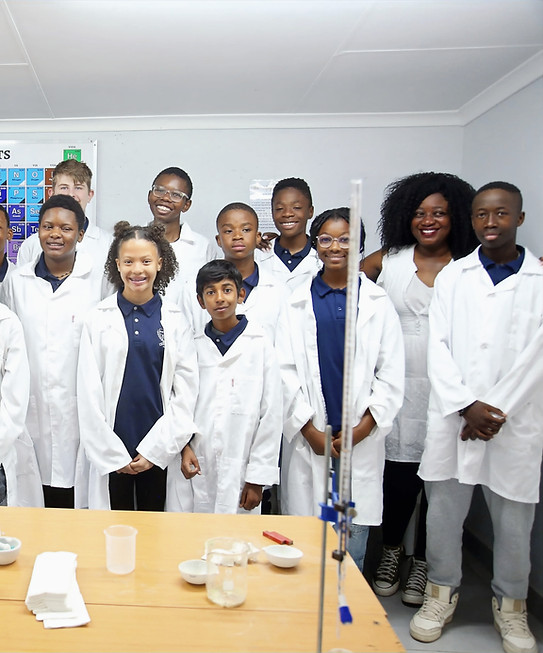 A diverse group of Midrand Montessori high school students in white lab coats standing with their science teacher in the laboratory.