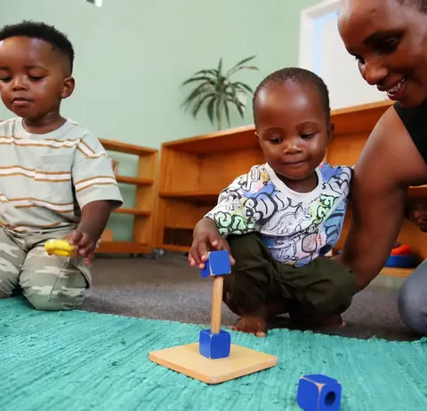 A toddler places a blue wooden cube onto a vertical wooden rod on a small base. A teacher kneels nearby, guiding gently, while another child plays with stacking rings in the background.