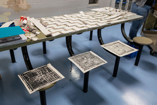 Three prints, depicting the dorm door ,sit on the stools attached to the table to dry, they are varying shades of black and gray because two of them are ghost prints