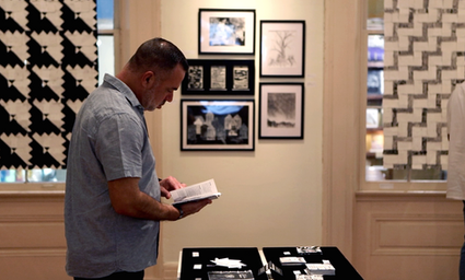 A person thoughtfully reads the book they picked up from among the others, behind them is a cluster of monochromatic artworks and blackout poems installed between the large quilt and weaving artworks suspended in tall windows 