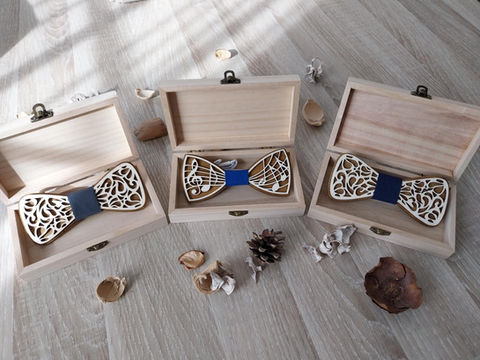 Trois nœuds papillon en bois gravés, présentés dans leurs boîtes en bois ouvertes, avec motifs décoratifs et tissu bleu marine au centre, entourés d’éléments naturels comme pommes de pin et coques végétales