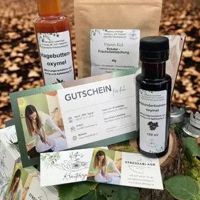 In einem herbstlichen Ambiente, umgeben von gefallenen Blättern, steht ein Geschenkset der „Stressablage“, das speziell zur Stärkung des Immunsystems zusammengestellt wurde. Im Vordergrund liegen mehrere hochwertige Gutscheinkarten der Stressablage, verziert mit natürlichen Illustrationen und einem Foto einer Behandlungsszene. Dahinter stehen drei Naturprodukte: eine Flasche Hagebutten-Oxymel, eine Flasche Holunderbeeren-Oxymel sowie eine Papiertüte mit dem Stressablagetee zum Stärken namens Vitamin Kick – Kräuter-Früchteteemischung. Die warmen Farben der Produkte harmonieren mit dem herbstlichen Hintergrund und betonen den stärkenden, wohltuenden Charakter dieses Sets.
