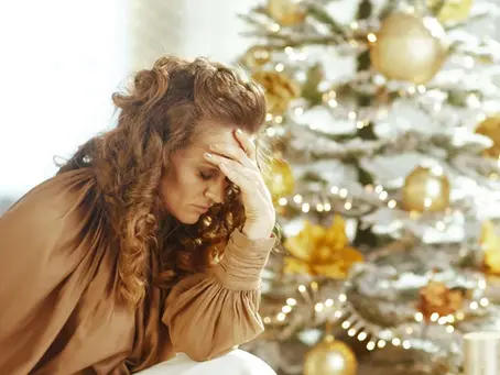Eine Frau mit langen Locken sitzt erschöpft vor einem weihnachtlich geschmückten Baum. Sie stützt den Kopf auf die Hand und wirkt gestresst, während goldene Kugeln und Lichter im Hintergrund glänzen.
