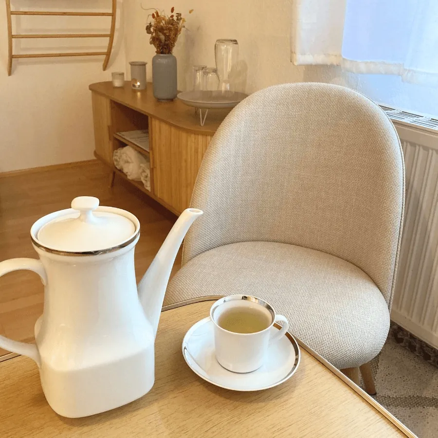 Einblick in die entspannte Atmosphäre der Stressablage in Steyr mit Regal und Sideboard aus Naturholz, grünen Pflanzen, naturfarbenem Sessel mit bequemem Stoffbezug, Kerzenschein, Getränkebar und Tee-Begleitung