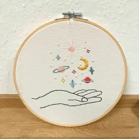 Im Shop der „Stressablage“ steht ein rund eingerahmtes Stickbild in einem Holzstickrahmen. Auf dem hellen Stoff ist eine stilisierte, schwarz gestickte Hand zu sehen, aus deren geöffneten Fingern bunte Himmelsmotive emporsteigen. Dazu gehören eine gelbe Mondsichel, kleine Sterne, ein pinker und ein blauer Planet, eine Sonne sowie funkelnde geometrische Sternformen in Rosa und Gelb. Die feine, sorgfältige Stickerei wirkt ruhig, liebevoll gestaltet und verleiht dem Bild eine symbolische, beinahe magische Ausstrahlung.