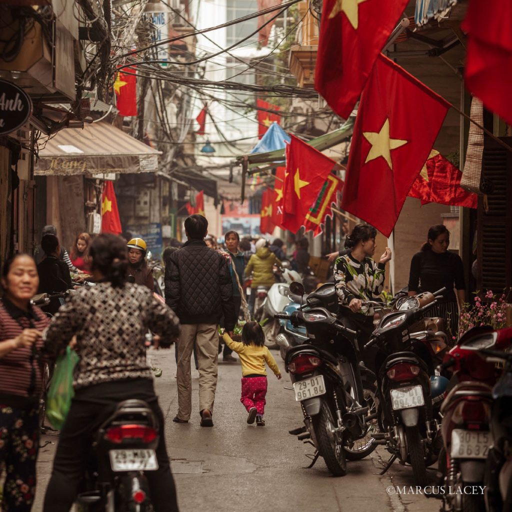 A lively street in Hanoi’s Old Quarter, adorned with red Vietnamese flags, with people walking, shopping, and motorbikes parked along the narrow alley.
