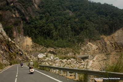 Vespa riders navigating a rugged mountain pass in Vietnam, surrounded by lush greenery and rocky cliffs.