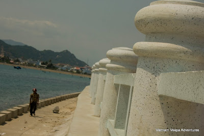 A coastal scene in Vietnam with a historic stone seawall, blue waters, and a traveler walking along the shore.