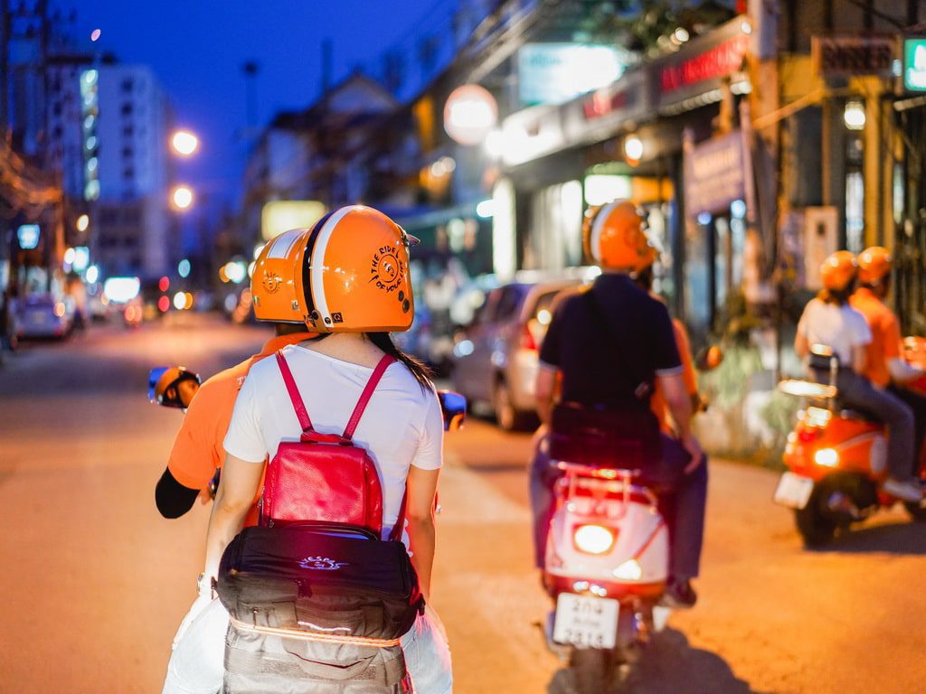 A group of Vespa riders wearing helmets navigating through the bustling streets of Chiang Mai at night.