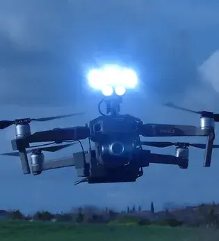 DJI mavic 2 drone with bright blue searchlights from Drone Sky Hook hovers over a grassy field under a cloudy sky, creating a futuristic and dynamic scene.