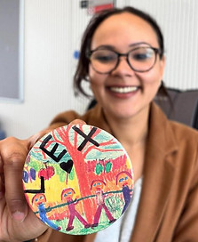 The image shows a person with glasses, who is slightly out of focus. They are smiling and holding up a handmade badge toward the camera. The badge has a multicoloured drawing on it with a tree in the background and in front of this, a line of people holding hands. At the top of the badge are the letters LEX in black, with a small heart.