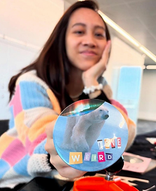 The image shows a person with long dark hair, their left hand resting on their chin. They are slightly out of focus but you can see they are smiling. They hold a handmade badge toward the camera. The badge has an image of a polar bear on it. Letters cut out from various magazines and articles have been cut out an arranged to spell 'love weird'.
