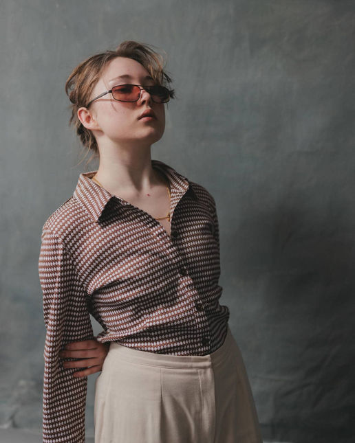 프레솔(Fressoul)**은 그녀가 직접 스타일링한 복고풍 의상을 입고 당당한 포즈를 취하고 있습니다. 크림색 다이아몬드 패턴 셔츠와 크림 베이지 코튼 바지, 브라운 안경으로 구성된 스타일은 부드러운 골드 체인을 더해 우아함을 강조합니다. 두 손을 뒤로 모은 채 옆을 바라보는 그녀의 모습은 런던 스튜디오의 어두운 회색 배경과 어우러져 품격 있는 분위기를 자아냅니다.