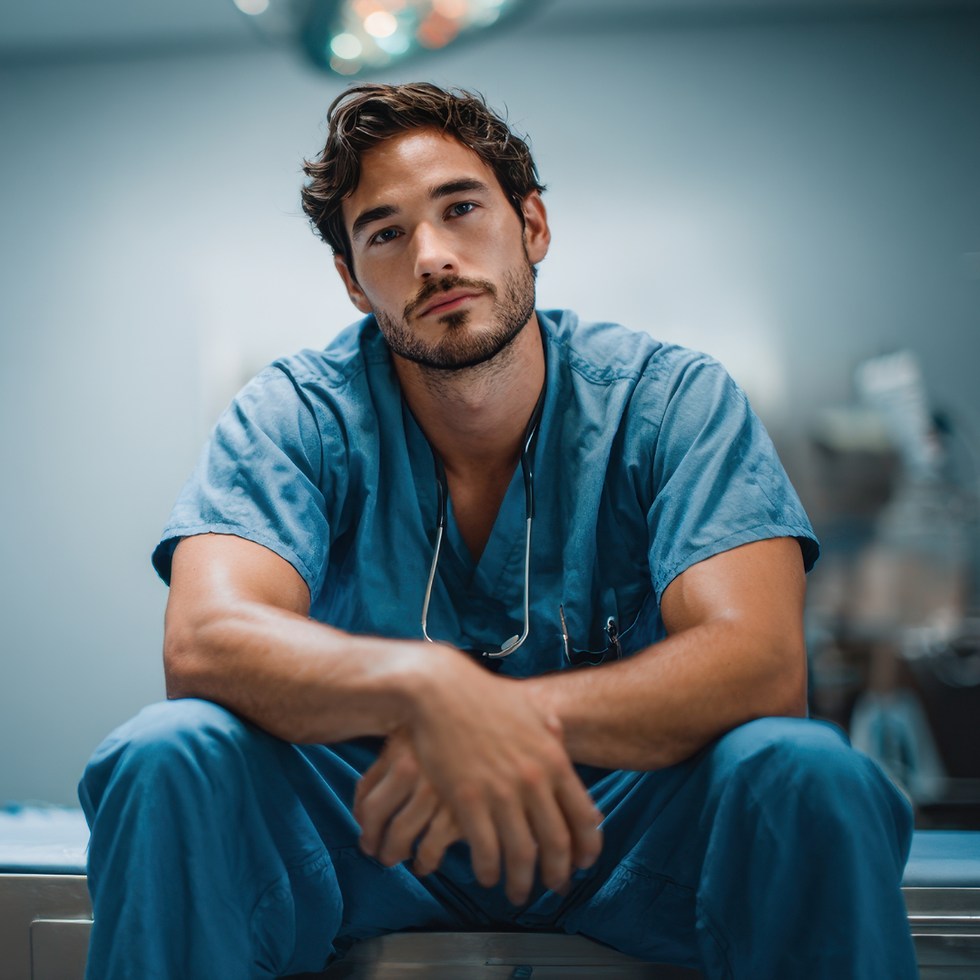 Man in blue scrubs sits on a hospital bed, looking thoughtful. Background is blurred, hinting at medical equipment, creating a calm mood.
