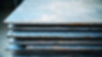 Close-up view of stacked steel plates showing edges with slight rust and wear, prepared for use in construction or manufacturing.