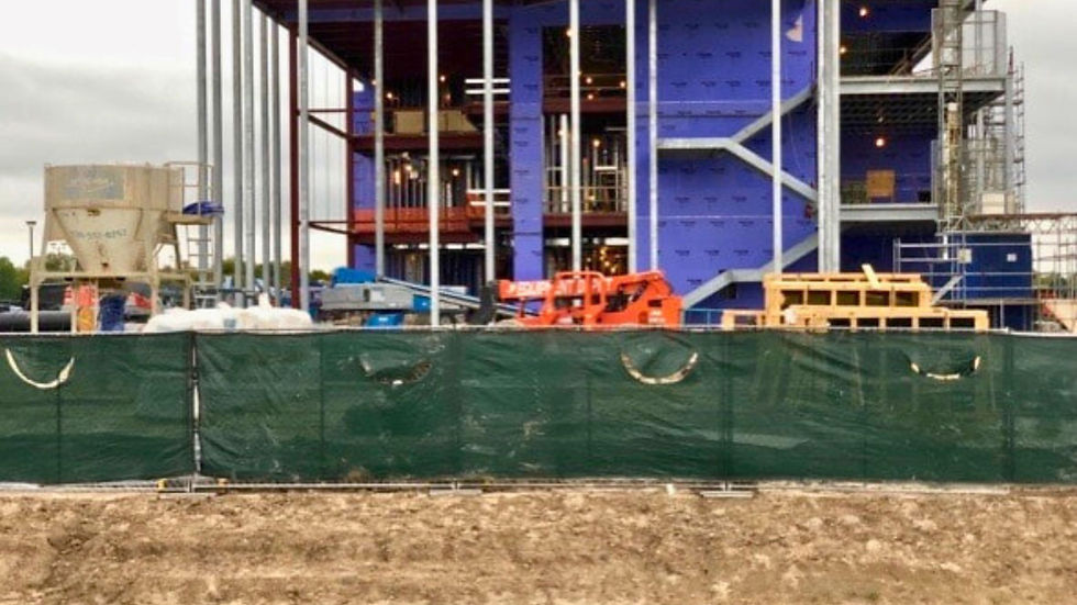 Construction site behind green privacy fencing, with metal wall studs and partial concrete structure visible above. A small piece of construction equipment is parked behind the fence, and the foreground features a landscaped roundabout with trimmed greenery.