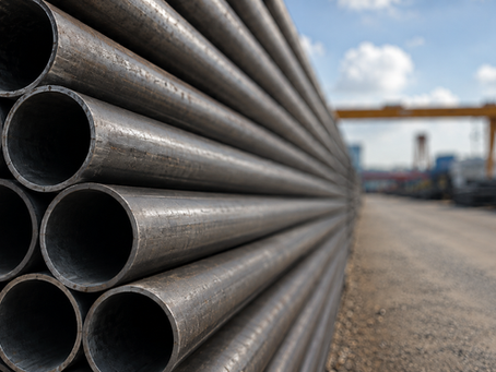 Close-up outdoor photo of stacked steel seamless pipes in an industrial yard. The pipes have a rugged, weathered look with light dust and surface wear, and the background shows a blurred gravel yard, blue sky, and industrial equipment.