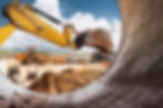 Close-up of a yellow excavator bucket on a construction site with dirt piles, a front loader, and building materials in the background.