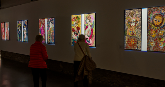 Visiteur en manteau rouge face à plusieurs portraits textiles accrochés, Katherine Roumanoff, exposition Trélazé Angers — Visitor in red coat facing multiple textile portraits, Katherine Roumanoff exhibition Trélazé