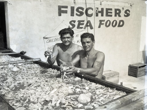 Two men holding shrimp behind a table covered in spring. A building behind has Fischer's Sea Food written on the side.