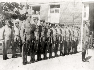 Rows of marines lined up outside a brick buildinghouse with red flowering bushes around it.