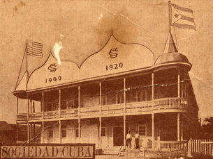 A building with a second floor gallery and cuban and american flags at each end with 1900 and 1920 written on the facade. Text on card says Sociedad Cuba.