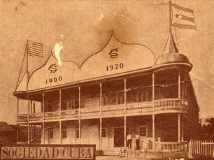 A building with a second floor gallery and cuban and american flags at each end with 1900 and 1920 written on the facade. Text on card says Sociedad Cuba.