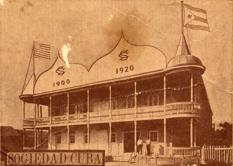 A building with a second floor gallery and cuban and american flags at each end with 1900 and 1920 written on the facade. Text on card says Sociedad Cuba.
