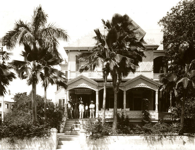 People standing on the porch of a two story house with palm trees in front of it.