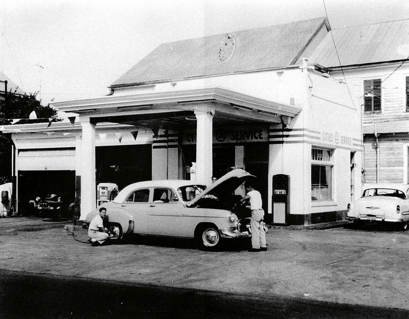Two men with a mid 20th century style car with its hood up in front of a service station.
