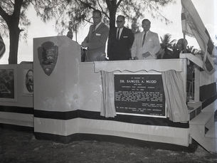 Three men at a podium with a National park Service shield and a plaque in memory of Dr. Samuel A Mudd.