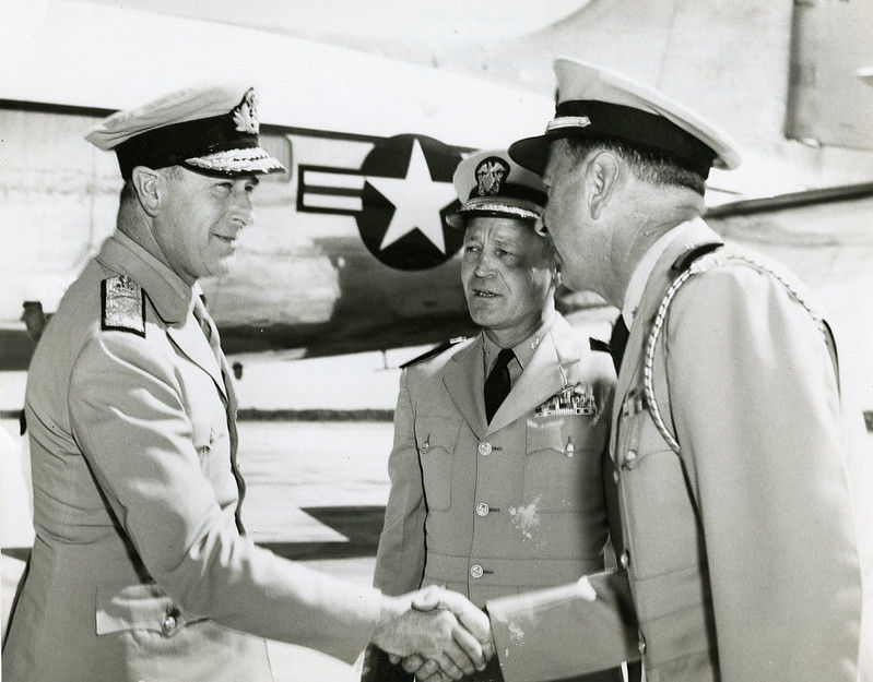 Two men in uniform shake hands in front of a third man in uniform