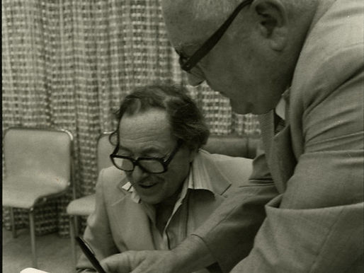 A man sits at a table while a man standing next to him points out drawings