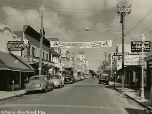 A commercial street with cars parked on the side and a banner that reads Americansim Week Junior Chamber of Commerce