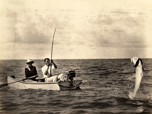 Two people in a small boat. One has a fishing rod with a large fish on the line.