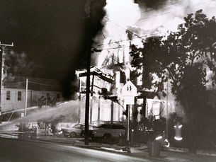 A firehose spraying water on a structure fully engulfed in flames