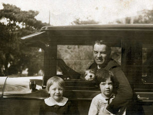 A man in a car with a dog on his lap and two children in front of the car