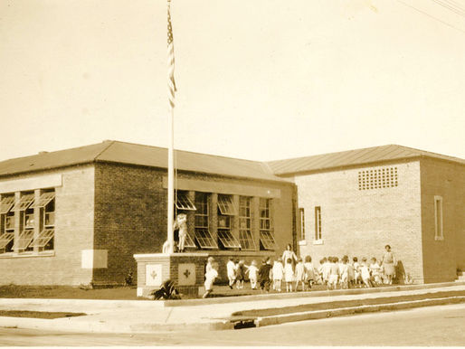 A brick building with a line of children, two adults, and a flagpole in the yard.