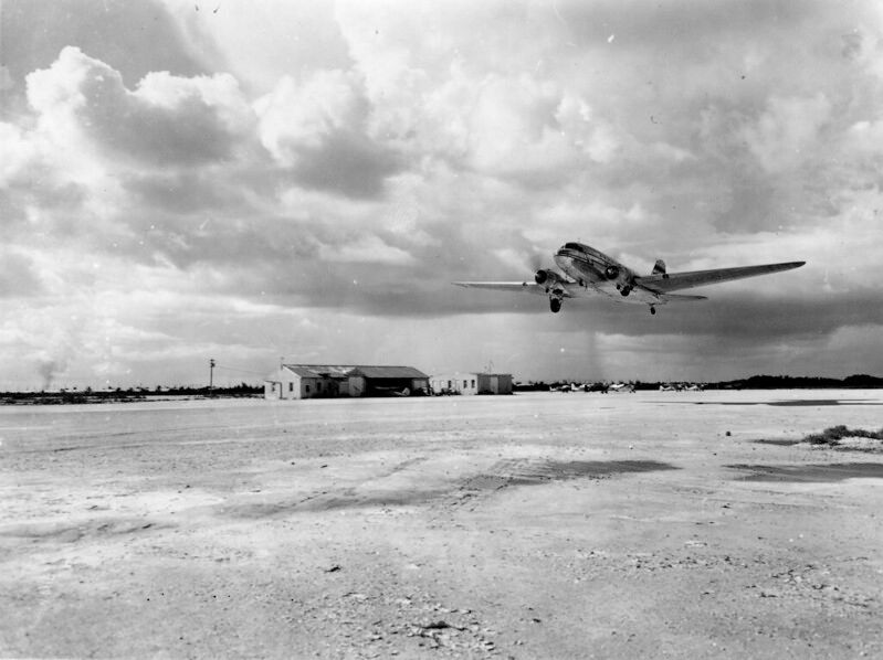 A propeller plane takes off from a field with two small buildings