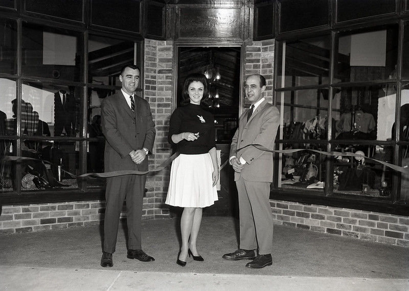 Three people in front of a storefront while the woman in the middle cuts a ribbon.