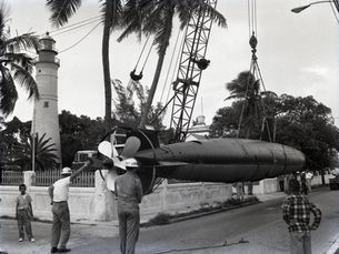 People standing around a submarine being suspended on a crane with a lighthouse in the background.