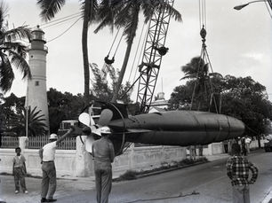 People standing around a submarine being suspended on a crane with a lighthouse in the background.