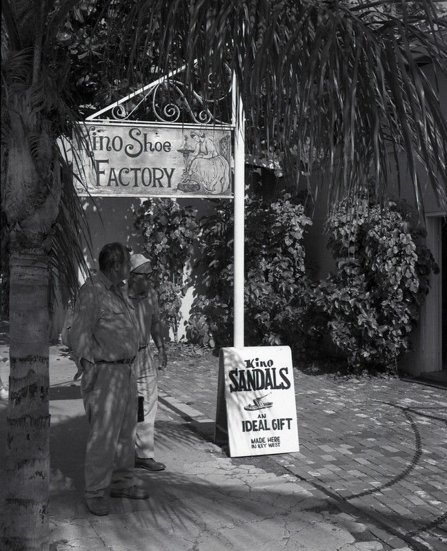 A man stands under a sign reading kino shoe factory with another sign on the ground reading kindo sandals make an ideal gift.
