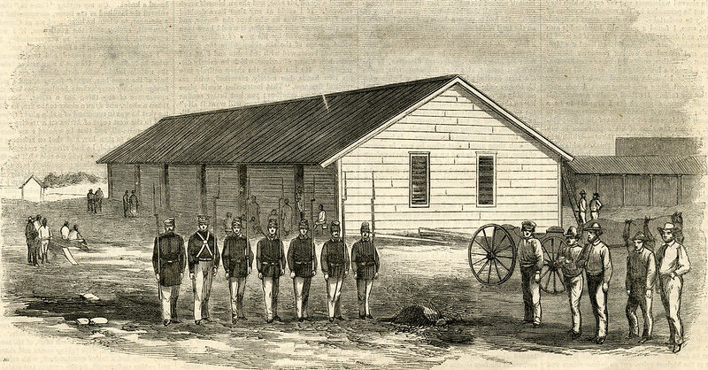 Soldiers in uniform stand in a line near a wooden building. A cannon and several people are in the background.