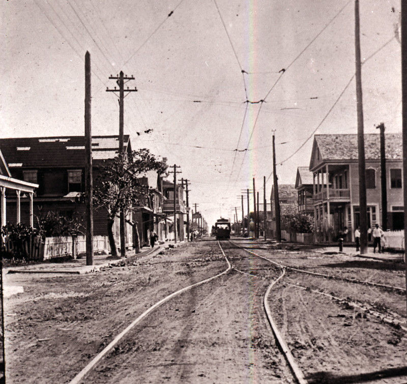 An unpaved street with buildings on the side and tracks and an electric trolley